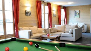 Una mesa de billar con bolas y tacos, un conjunto de sofás y una mesa de billar en una habitación con paredes de madera y ventanas grandes con cortinas rojas.