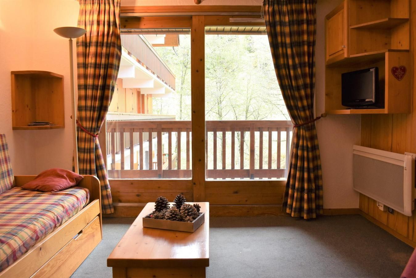 A cozy cabin room with a bed, a small table with pinecones, a TV, and a balcony with a wooden railing.