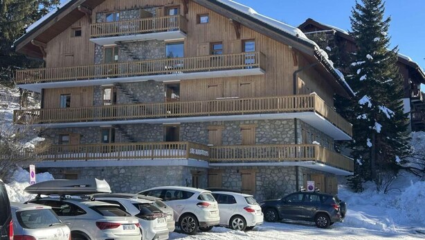 A multi-story wooden building with balconies, a stone facade, and a parking area with cars.