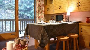 A cozy dining area with a table set for two, wooden cabinets, and a view of a forest through a large window.