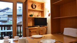 Un comedor de madera con una mesa preparada para dos, un televisor sobre un soporte de madera y vistas exteriores a una casa de madera.