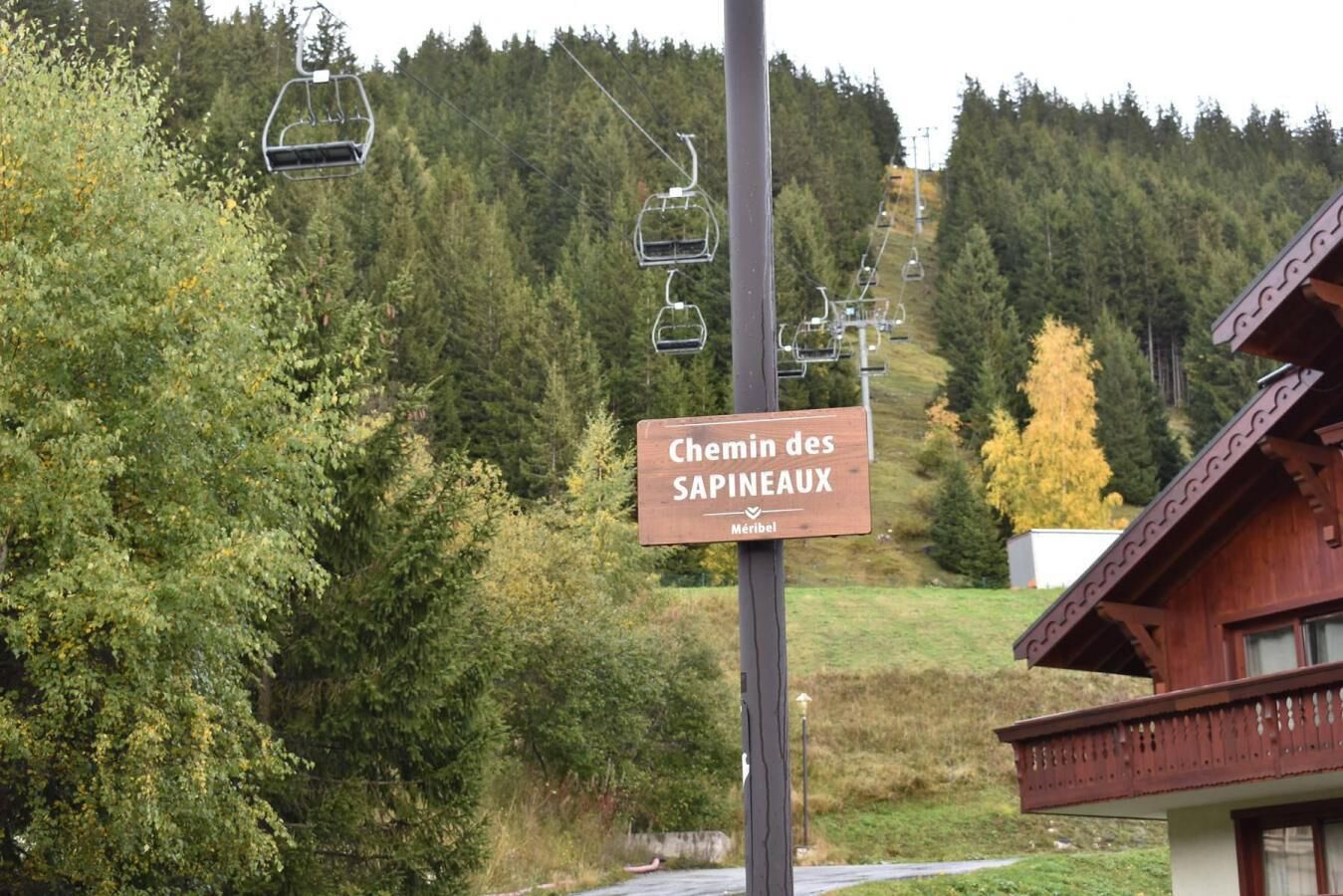 A sign for 'Chemin des SAPINEAUX' with a wooden post, a mountain backdrop, and a ski lift in the distance.