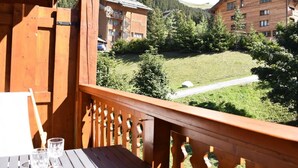 Un balcón de madera con una mesa y dos vasos, con vistas a una zona montañosa con edificios y vegetación.