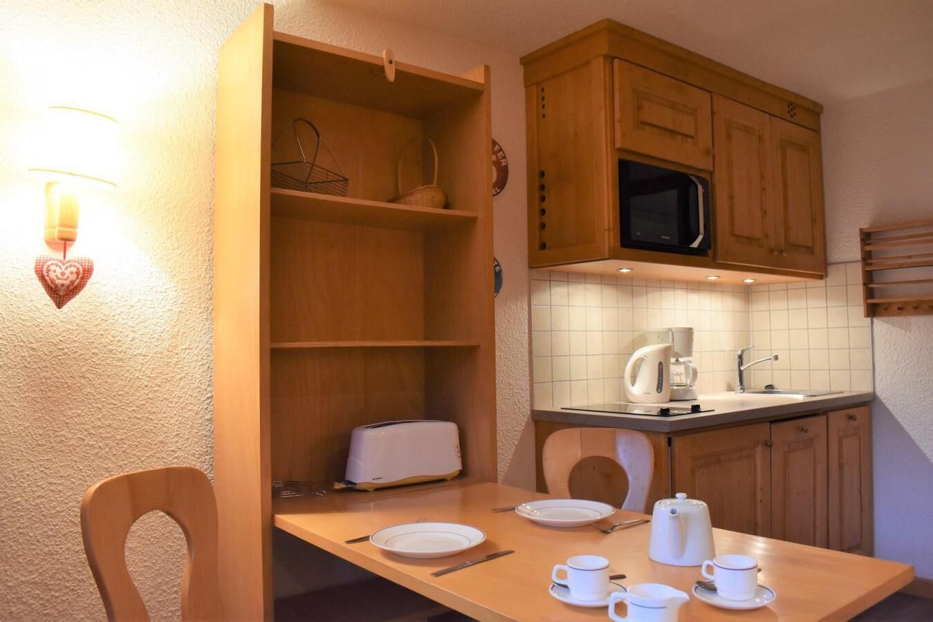 Een kleine keuken met houten kastjes, een magnetron, een spoelbak en een eettafel gedekt met borden, kopjes en een waterkoker.