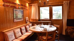 Un accogliente ambiente in legno con un tavolo rotondo per quattro persone, una panca con cuscini a motivi floreali e un'ampia finestra con tende.