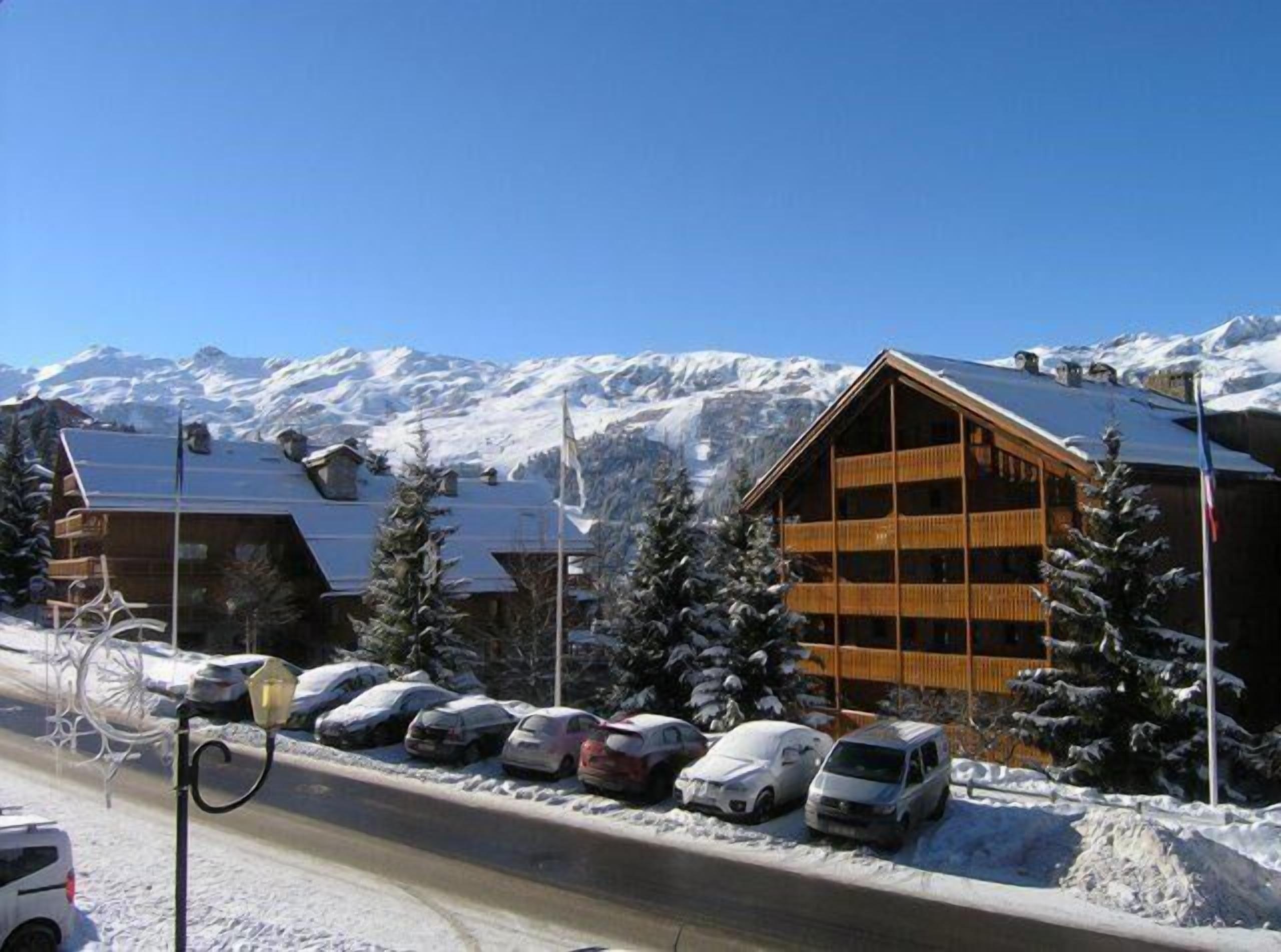 A wooden lodge with multiple balconies, a parking area with several cars, and a backdrop of snow-covered mountains.