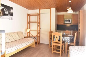 A compact room with a wooden ceiling, a sofa with patterned upholstery, a dining table with chairs, and a kitchenette with a microwave.