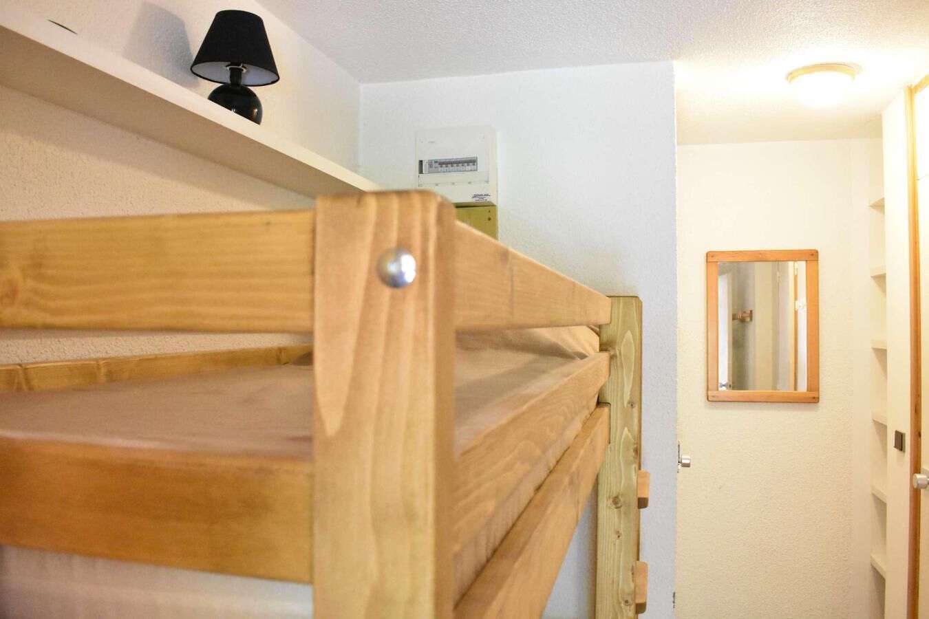 Una cama litera con estructura de madera y un tornillo metálico, una lámpara negra en un estante y un termostato fijado a la pared.