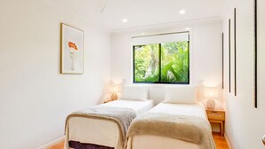 A room with two beds, a window with a view of greenery, a framed picture on the wall, and bedside lamps.