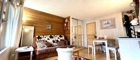 Een gezellige woonkamer met een houten wand, een bank, een witte fauteuil, een eettafel en een keukenhoek.