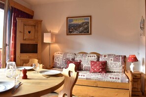 A cozy room with a wooden dining table set for two, a sofa with patterned cushions, a cabinet, and a wall-mounted lamp.