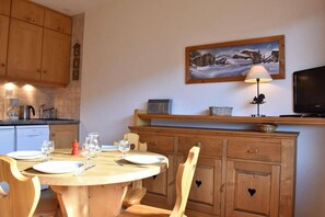 A wooden dining table with chairs, a kitchen area with cabinets, a painting, a lamp, and a television.