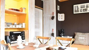 A dining area with a wooden table set for two, a coffee machine, and a shelf with dishes and cups.