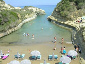 A beach with clear waters, rocky cliffs, and several sun loungers and umbrellas. - Livadi Villa (Poulimates)
