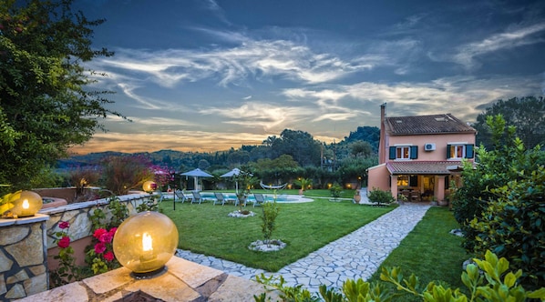 A well-maintained garden with a swimming pool, stone pathway, and a house with a tiled roof. - Livadi Villa (Poulimates)