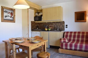 Une cuisine compacte avec des meubles en bois, un four à micro-ondes et un évier. On y trouve une table à manger dressée avec des assiettes et des verres, ainsi qu’un canapé à carreaux dans l’espace salon.