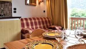 Un coin repas chaleureux avec une table en bois dressée pour deux personnes, un canapé à carreaux et une vue sur la verdure environnante.