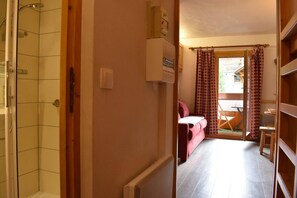 A narrow hallway with a shower on the left, a wooden door leading to a room with a pink sofa and a table, and a window with patterned curtains. - 300m from shops, 1st floor, balcony, tv, ski locker, 18m², Méribel (Méribel - Les Allues)