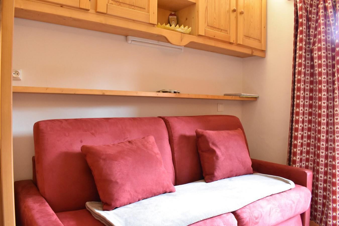 A red sofa with two pillows and a white blanket, wooden cabinets above, and a patterned curtain on the right.