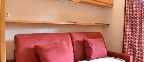 A red sofa with two pillows and a white blanket, wooden cabinets above, and a patterned curtain on the right.