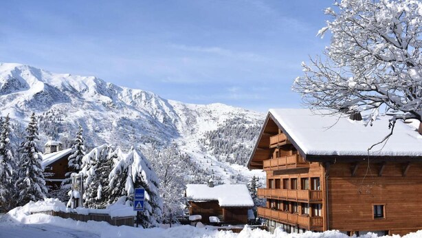 A wooden lodge with a snow-covered roof, surrounded by snow-covered trees and mountains.