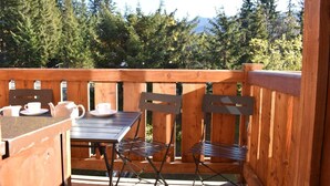 A wooden deck with a table and chairs, a small table with a teapot and cups, and a view of trees and mountains.