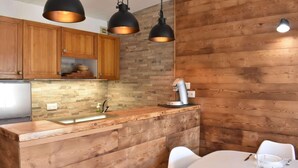 A kitchen with wooden cabinetry, a stone backsplash, and a built-in island with a sink. There are pendant lights above the island and a coffee machine on the countertop.