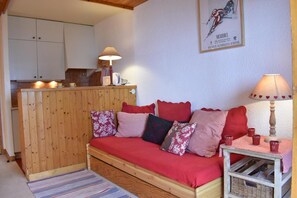 Una habitación acogedora con un sofá de madera, una mesita con una lámpara y una zona de cocina al fondo.