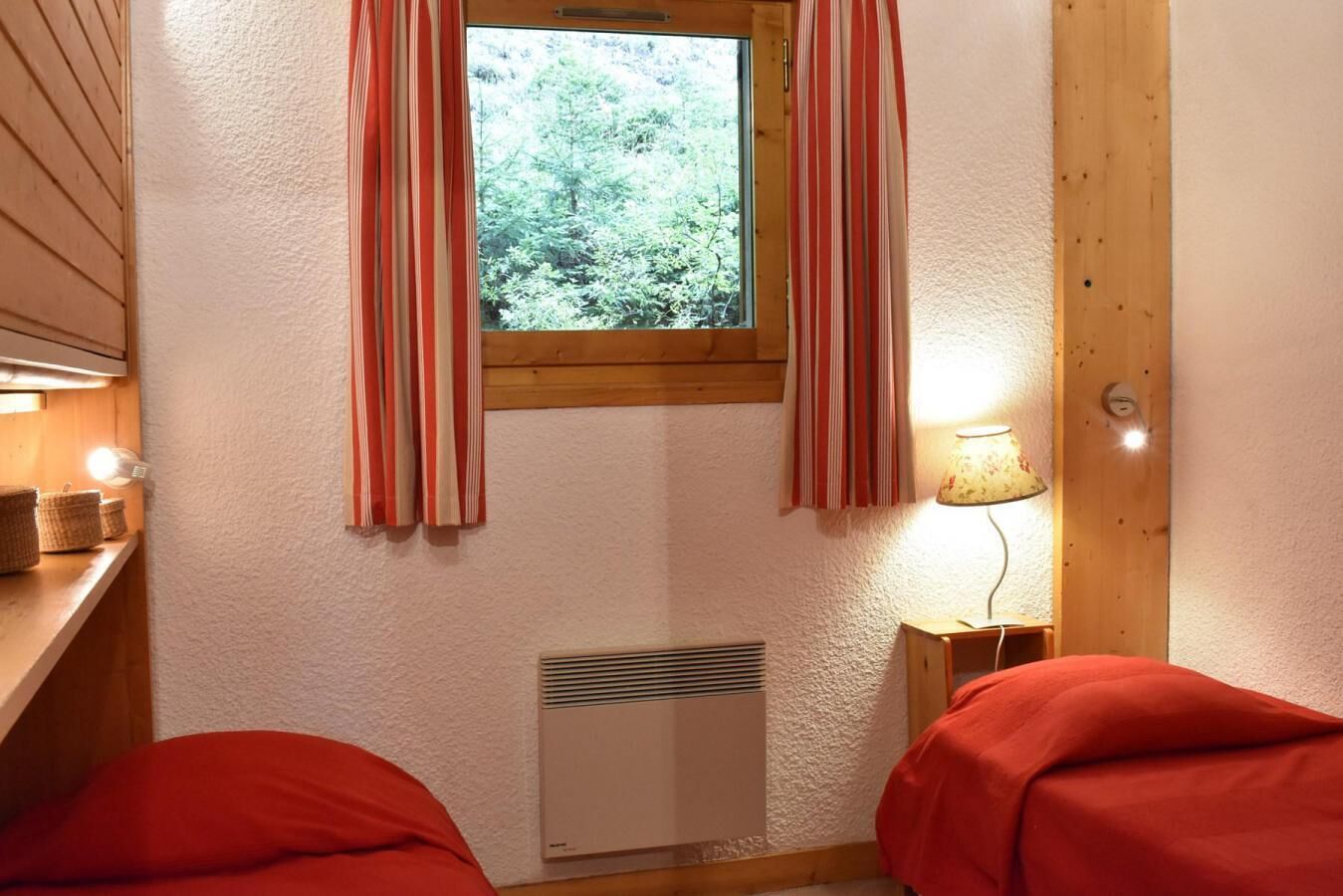 A room with two beds, a window with curtains, a wall-mounted heater, and a lamp on a side table.