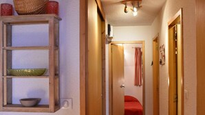 Un pasillo estrecho con un mueble de madera en la pared izquierda, una puerta que da a una habitación con colcha roja, y una lámpara en el techo.
