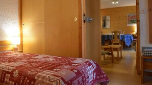 Une chambre d’hôtel avec un lit, une porte en bois, une petite table et un coin repas visible à travers une porte ouverte.