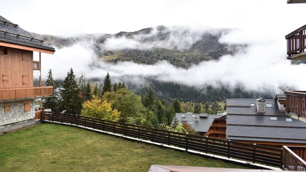 Un chalet de montagne doté d’une terrasse en bois et offrant une vue sur une vallée boisée et des montagnes.