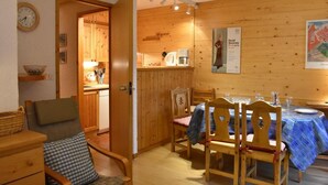 Une pièce chaleureuse aux murs en bois, une table à manger dressée pour quatre personnes et un coin cuisine visible à travers une porte ouverte.