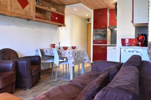 A cozy living room with a sofa and chairs, a dining table with a tablecloth, and a kitchen area with red cabinets and appliances.