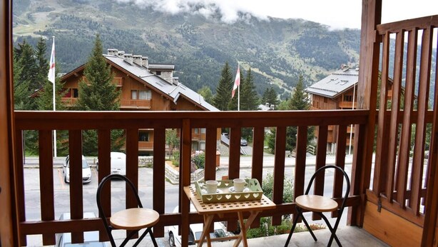 Ein Balkon mit zwei Stühlen und einem Tisch, mit Blick auf eine Berglandschaft mit Gebäuden und Flaggen.