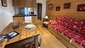 Une pièce chaleureuse avec une table à manger en bois, dressée pour deux personnes, un canapé rouge à motifs et une kitchenette dotée de meubles en bois et d’un réfrigérateur en acier inoxydable.