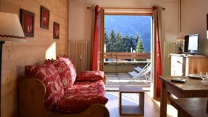 Une pièce chaleureuse avec un décor intérieur en bois, un canapé rouge à motifs, une petite table avec un verre, et une vue sur les arbres par la fenêtre.