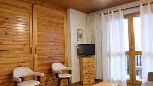 A cozy room with wooden walls, a television on a wooden cabinet, two white chairs, a wooden table, and a large window with curtains.