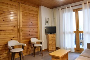 Una stanza accogliente con pareti in legno, una televisione su un mobile in legno, due sedie bianche, un tavolo in legno e un'ampia finestra con tende.