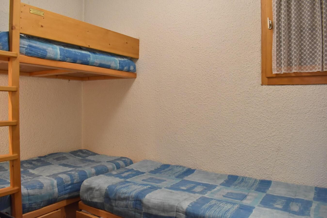 A bunk bed room with a wooden ladder, two beds with blue and white patterned bedding, and a window with a patterned curtain.
