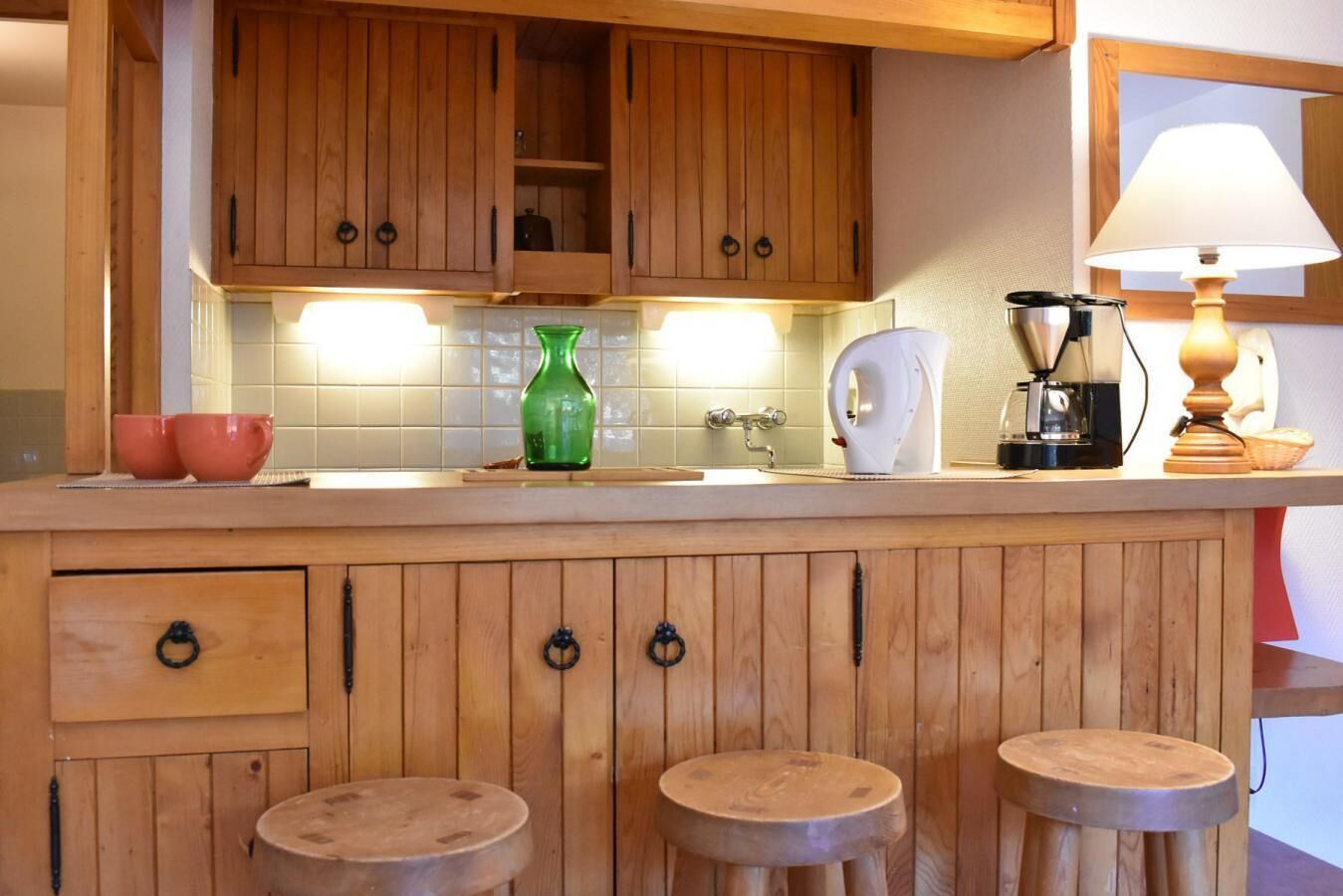 Une cuisine avec des meubles de rangement en bois, un plan de travail, un vase vert, une machine à café et une lampe.