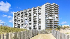 Un edificio residenziale a più piani con balconi, un vialetto sabbioso che conduce all'ingresso e una recinzione in legno.