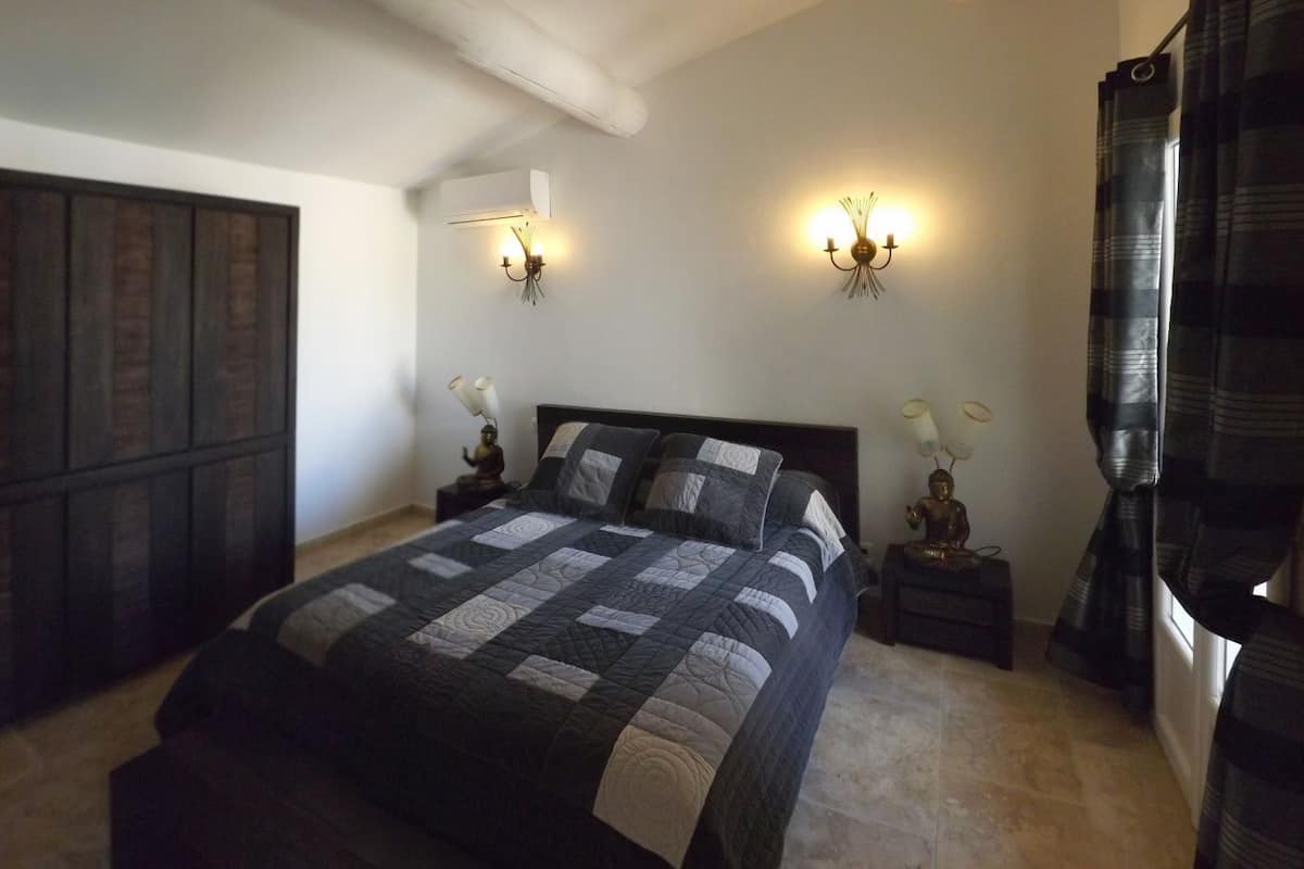A bedroom with a large bed, two wall sconces, a nightstand with a lamp, a dark wooden wardrobe, and a window with curtains.