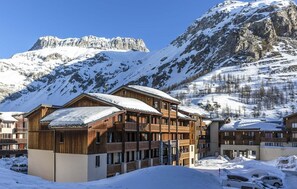 Ein Chalet-Hotel aus Holz mit mehreren Balkonen, vor einer Kulisse schneebedeckter Berge und blauem Himmel.