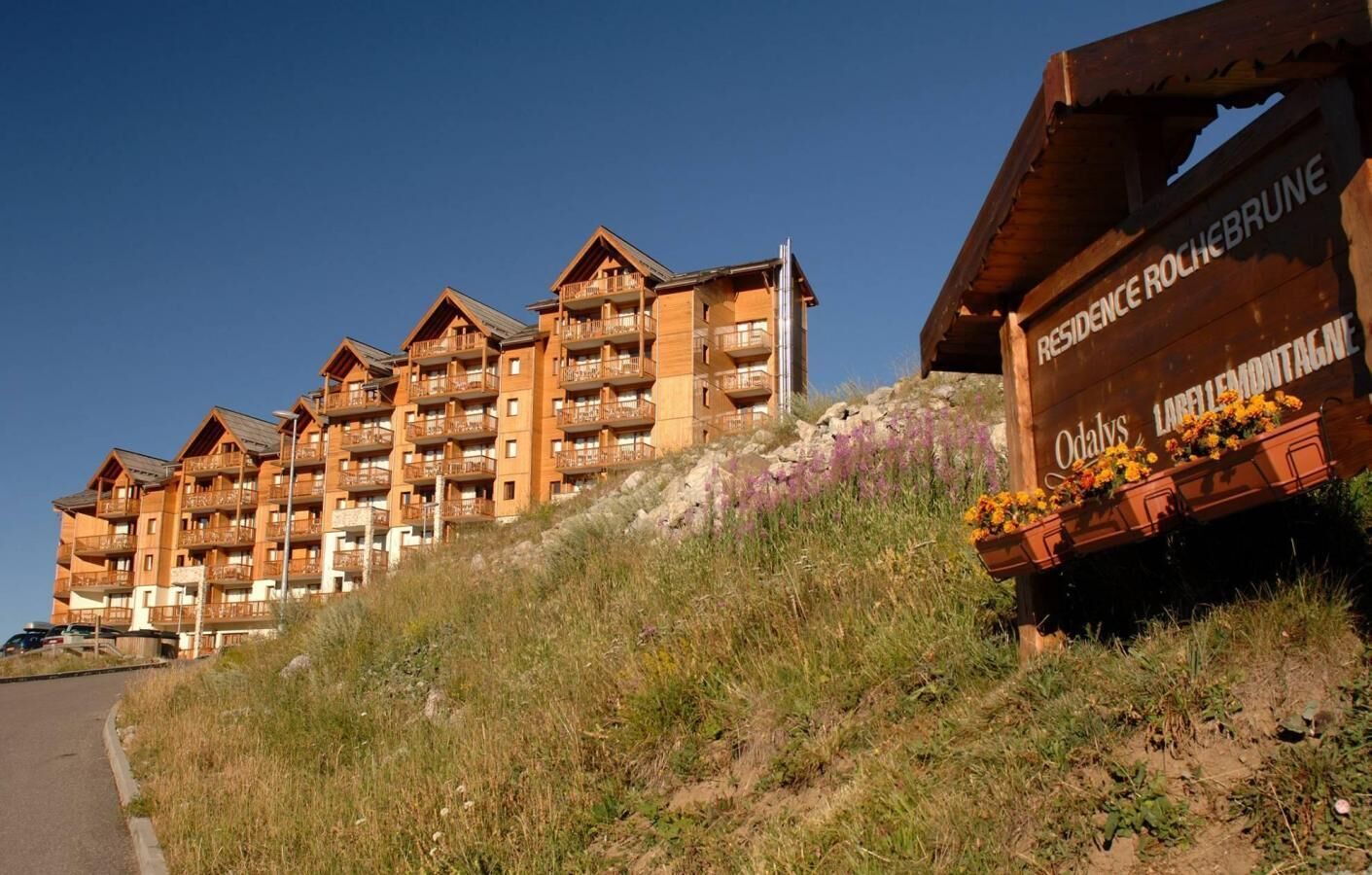 Un hôtel à plusieurs étages doté de balcons, une enseigne indiquant « Résidence Rochebrune » et un parterre de fleurs aux fleurs jaunes.