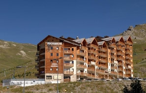 A multi-story hotel with a wooden facade, balconies on each floor, and a mountainous backdrop.
