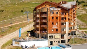 A multi-story hotel with wooden exterior and balconies, a swimming pool, and ski lift infrastructure in the background. - Ski-in/ski-out & foot of the slopes, swimming pool, balcony, tv, 40-45m², Orcières Merlette 1850 (Orcières)