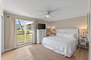 A bedroom with a bed, a TV, a ceiling fan, a lamp, and a view of a grassy area outside. - 2BR Condo on Golf Course w/ Free Beach Service (Destin)