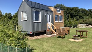 A small, two-story house with a grey exterior and a wooden upper floor. It features a small deck with outdoor furniture and a fenced garden area. - The Cedar Tiny House (Coldingham)