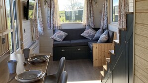 A cozy cabin interior with a wooden ceiling, a sofa with floral cushions, a dining table with chairs, and a window with curtains. - The Cedar Tiny House (Coldingham)